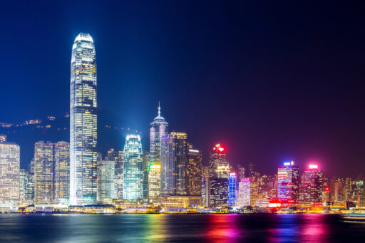 Colorful nighttime view of the Hong Kong skyline with illuminated skyscrapers reflecting on Victoria Harbour, evoking the setting of High Rise by Harper Bliss.